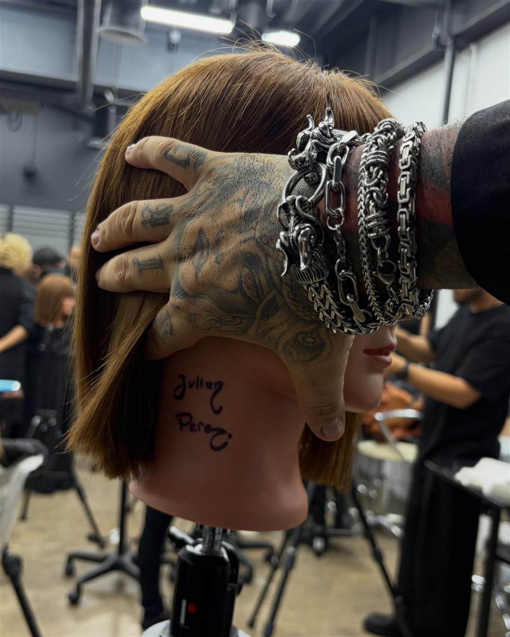 Mano tatuada con pulseras de plata sosteniendo un corte bob en una cabeza de práctica